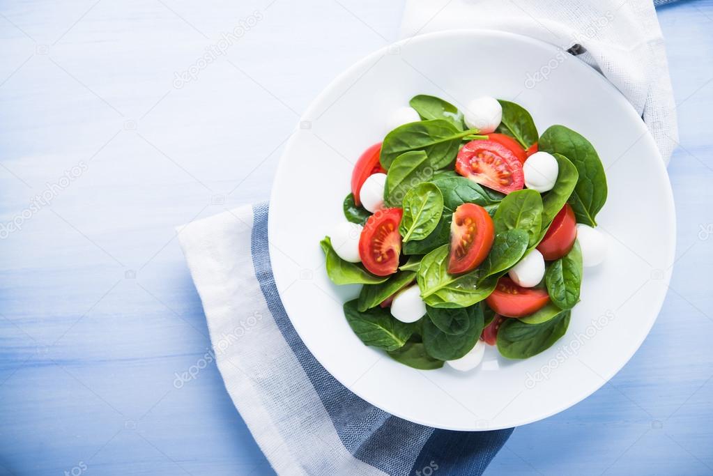 Fresh salad with mozzarella cheese, tomatoes and spinach on blue wooden background top view.