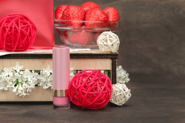 Damgasız pembe ruj, kasede taze çilek, rattan topları, beyaz leylak çiçekleri ve koyu ahşap arka planda kırmızı kozmetik çanta. Kozmetik ürünler, dudak kremi. Markalaşma kavramı