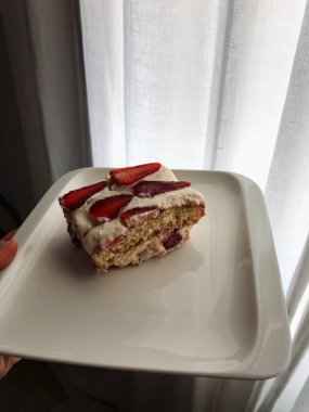 Pencere yakınındaki Beyaz Tabakta Çilek Dilimli Tiramisu Sardunya Evinde Dikey Yakın Çekim