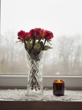 Vazodaki gül buketi ve Beyaz Örgü Peçetedeki Parlak Mum Pencere kenarındaki Çıplak Ağaçlar ve Gri Gökyüzü Puslu Görüş
