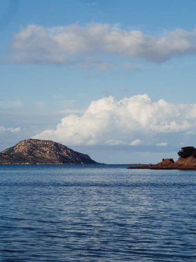 Deniz, tepeler ve mavi gökyüzünde kabarık beyaz bulutlar ile dikey Sardunya deniz manzarası suya, Corallina plajına, Porto San Paolo, İtalya 'ya yansımaktadır.