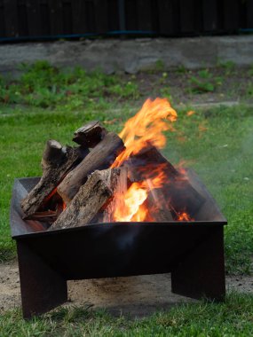 Çağdaş kare şeklindeki, bacakları olan metal bir ateş çukurunda odun yakmak, parlak alevler sıcak bir atmosfer yaratır. Yeşil çimenlikte modern arka bahçe detayı, insan yok, dikey yakın plan..