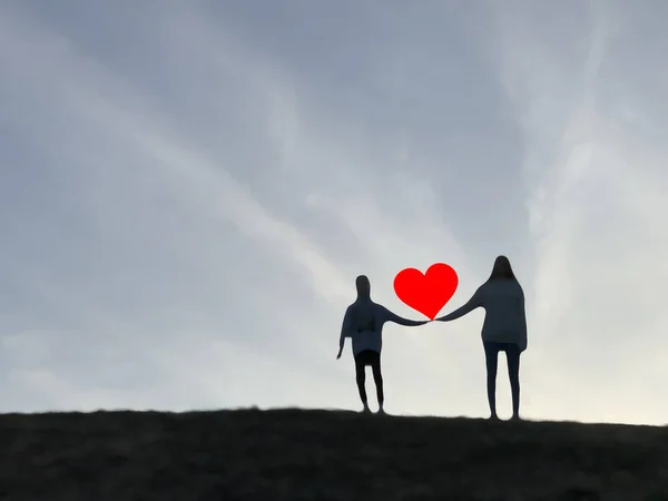 Silhouette of two people on top of the mountain. A loving couple with a heart in their hands, against the background of the sky. The guy and the girl are holding a red heart.