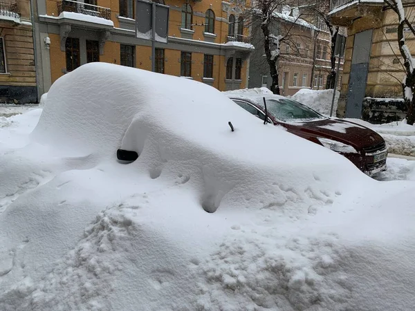 The car was covered with snow in the parking lot. Cars under a large layer of snow. Snowfall on the streets of a European city.