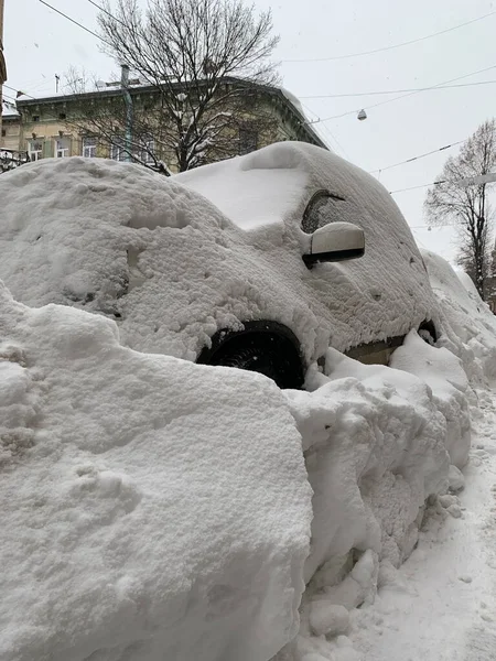 The car was covered with snow in the parking lot. Cars under a large layer of snow. Snowfall on the streets of a European city.