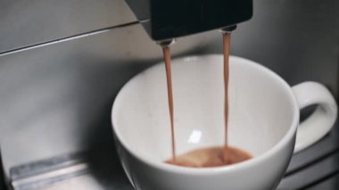 Kahve makinesinden taze demlenmiş kahve beyaz bir seramik fincana dökülüyor.