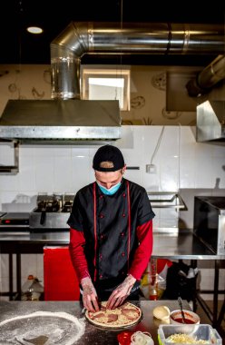 Şef koruyucu maske takıyor, mutfakta pizza yapıyor, yemek pişiriyor. 