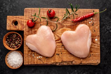 Tuz, biber, sarımsak ve koyu arkaplanda taze otlar bulunan kalp şeklinde çiğ tavuk eti.