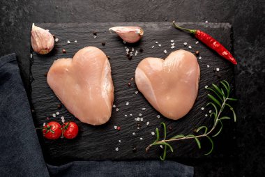 Tuz, biber, sarımsak ve koyu arkaplanda taze otlar bulunan kalp şeklinde çiğ tavuk eti.