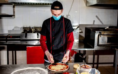 Şef koruyucu maske takıyor, mutfakta pizza yapıyor, yemek pişiriyor. 