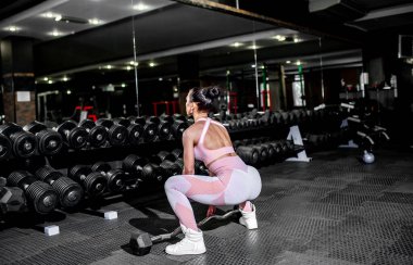 Spor salonunda dambıllarla egzersiz yapan genç bir kadın.