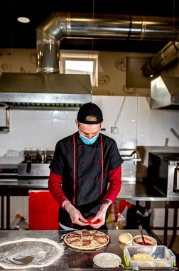 Şef koruyucu maske takıyor, mutfakta pizza yapıyor, yemek pişiriyor. 