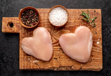 Tuz, biber, sarımsak ve koyu arkaplanda taze otlar bulunan kalp şeklinde çiğ tavuk eti.