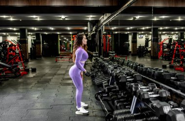 Spor salonunda dambıllarla egzersiz yapan genç bir kadın.