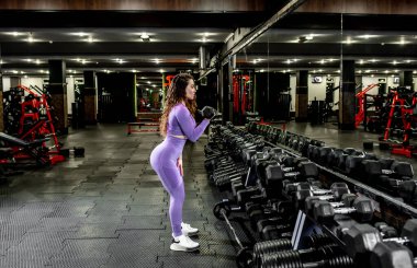Spor salonunda dambıllarla egzersiz yapan genç bir kadın.