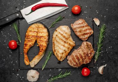 Siyah yüzeydeki malzemelerle çevrili ızgara balık ve et bifteği.