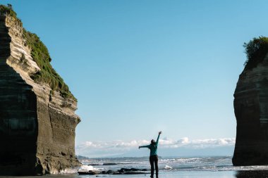 Kadın sahildeki iki kaya oluşumunun ortasında kollarını açıyor. Seyahat kavramı.