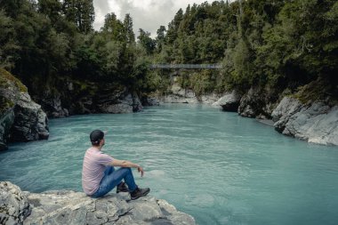 Genç adam bir kayanın üzerinde oturuyor ve Hokitika Gorge, Yeni Zelanda, Güney Adası 'ndaki asma köprüye bakıyor..