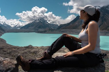 Esmer kadın bir kayanın üzerinde dinleniyor ve Tasman Gölü ve Atına bakıyor. Yeni Zelanda, Güney Adası
