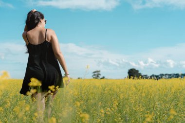 Kırsalda sarı çiçek tarlalarının arasında yürüyen esmer bir kadın. Gerçek İnsanlar konsepti