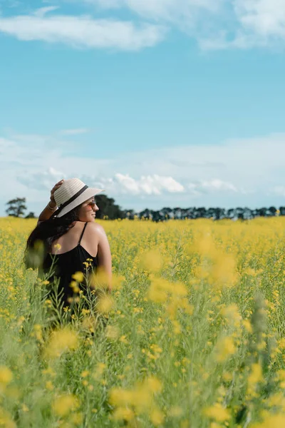 Çiçeklerle dolu sarı tarlada profilden poz veren yaz şapkası takan genç bir kadın. Dikey fotoğrafçılık. Gerçek İnsanlar konsepti