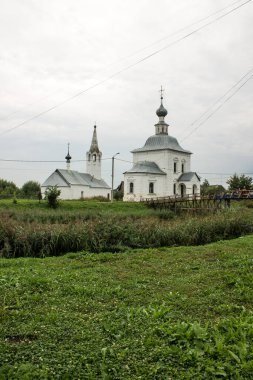 SUZDAL, Vladimir Bölgesi, RUSSIA-AUGust, 11, 2021: Epifani ve Noel beyaz kiliseleri bulutlu bir gökyüzüne karşı yeşil çimlerde ve kopyalamak için bir alan