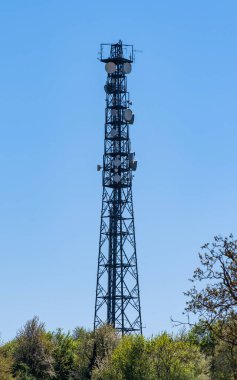 Telekomünikasyon direği, kablosuz TV antenleri yeşil çitlerin üzerindeki mavi bulutsuz gökyüzüne karşı