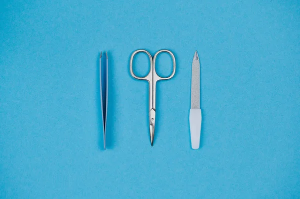 Sfondo blu, al centro si trovano pinzette sdraiate, lima per unghie e forbici per unghie — Foto Stock