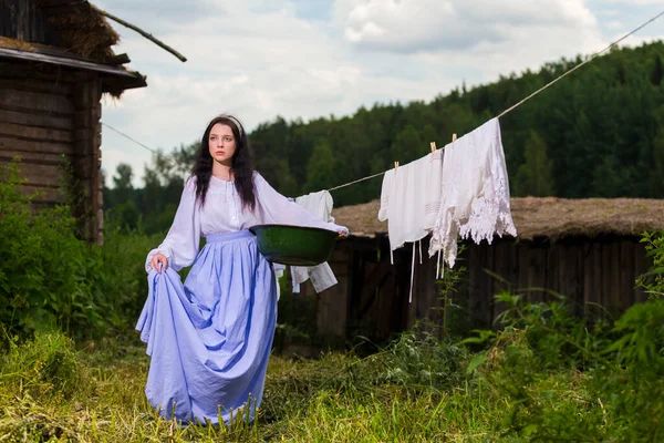 Portrait of Caucasian Brunette Girl in Traditional Rural Dress Holding Basin With Linens in Countryside. Horizontal Image