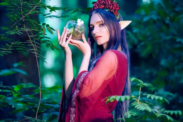 Portrait of Caucasian Girl With Artistic Elf Ears and Flowery Garland Holding Glass Dome With Grounded Flower in Summer Forest. Horizontal Image