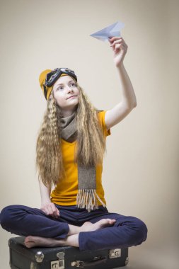 Uçan Gözlüklü Genç Kız, eski seyahat çantasının üzerinde otururken Origami Kağıt Uçakla Poz Veriyor. Dikey Resim Kompozisyonu