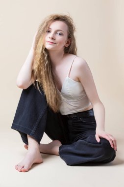 Beyaz sutyenli, kot pantolonlu, bej arka planda katlanmış bacaklı, beyaz, genç, sakin sarışın kız. Dikey Resim Yönlendirmesi