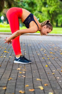 İleriye doğru esneme egzersizleri sırasında gündüz vakti park alanında vücut bükme egzersizleri yapan beyaz bir kadın. Dikey Resim