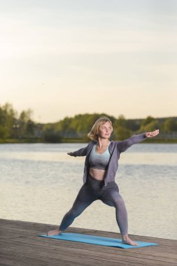 Yoga yaparken, dışarıda, tahta sahnenin üzerinde gerinen olgun, beyaz bir kadın. Dikey Resim Kompozisyonu