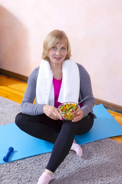 Smiling Mature Woman Having Break During Home Fitness Exercises Holding Glass Bowl Of Fresh Vegetables. Vertical Shot