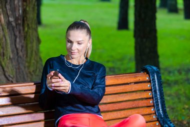 Açık havada müzik çalanlarla antrenman yaparken spor kıyafetleri içinde dinlenen beyaz kadın koşucunun yakın çekimi. Yatay Resim Kompozisyonu