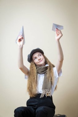 Beyaz üniformalı genç bir kız ve bej arka planda çantanın üzerinde otururken Origami kağıt uçaklarıyla sokak kepi pozu veriyor. Dikey Resim Yönlendirmesi