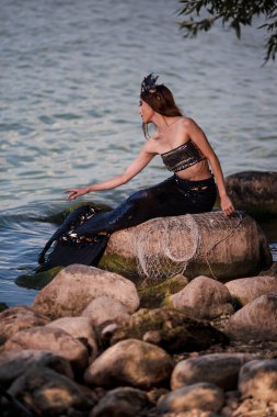 Kayalıklarda Deniz Kıyısında Ağlı Asyalı Denizkızı. Deniz kabuğu desenli Taç ve Denizin Sahibe Kraliçesi kılığında siyah parlak kuyruk takıyor. Dikey resim