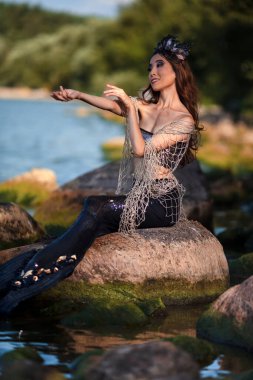 Deniz kıyısındaki Kayalıklarda Deniz Kızları 'nın özlemi. Deniz kabuğu desenli taç takıyor ve denizin kraliçesi gibi sert vücutla kaplı siyah parlak kuyruk takıyor. Dikey Resim