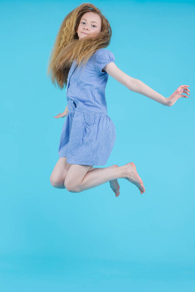 Happy Jumping Teenager Blond Girl in Long Blue Dress Against Blue Background. Vertical Image Composition