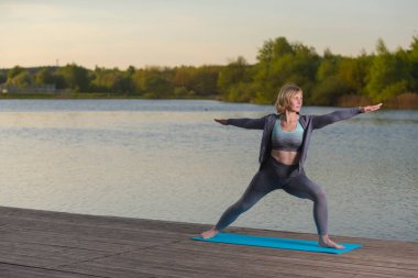 Spor Konseptleri. Yoga yaparken, dışarıda, tahta sahnenin üzerinde gerinen olgun, beyaz bir kadın. Yatay Resim Kompozisyonu