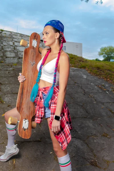 Hipster Kaykaycı Grunge Kiremitleri 'nde uzun kaykaylı poz verirken kaykaylı ve güneş gözlüklü şapka takıyor. Dikey Resim