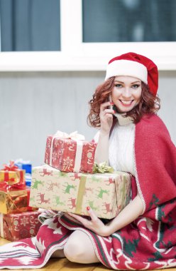 Happy and Laughing Caucasian Santa Girl with Lots of Presents Behind. Talking by Cellphone and Sitting Under the Plaid Indoors