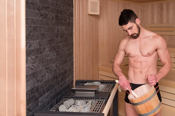 Schöner Mann gießt Wasser auf heißen Felsen in der Sauna — Stockfoto