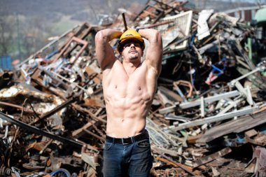 Endüstriyel hurdalıkta dikilen ve iyi eğitilmiş vücudunu sarı kaskla gösteren genç bir adamın portresi.