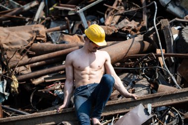 Endüstriyel hurdalıkta oturan ve iyi eğitilmiş vücudunu sarı kaskla gösteren genç bir adamın portresi.