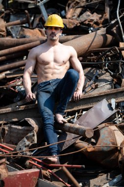 Eski Hurda Metal Garajında ve Esnek Kaslarda Güçlü Oturan Genç Adam - Kaslı Atletik Vücut Geliştirici Spor Modeli Sarı Miğferli Pozisyon