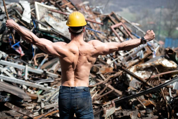 Young Man Standing Strong in Old Scrap Metal Garage and Flexing Muscles - Muscular Athletic Bodybuilder Fitness Model Posing With Yellow Helmet