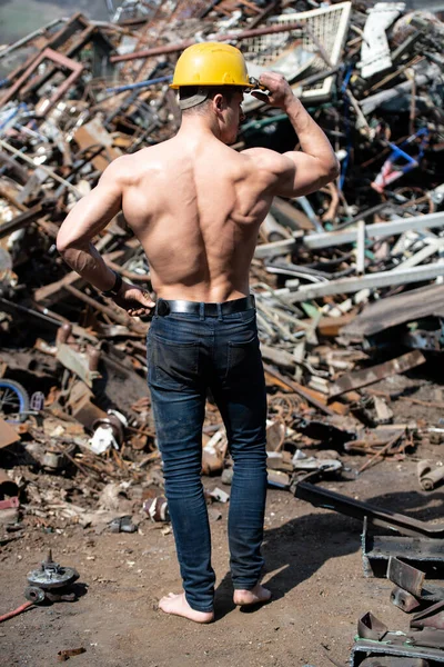 Handsome Man Standing Strong in Scrap Metal Industrial Junkyard and Flexing Muscles - Muscular Athletic Bodybuilder Fitness Model Posing With Yellow Helmet