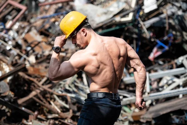 Handsome Man Standing Strong in Scrap Metal Industrial Junkyard and Flexing Muscles - Muscular Athletic Bodybuilder Fitness Model Posing With Yellow Helmet